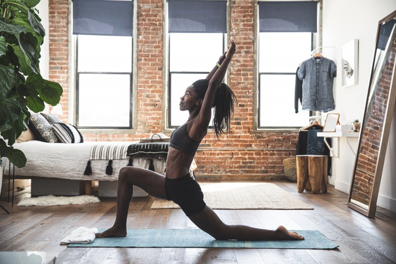 Young woman in gray yoga clothes doing yoga in her bedroom with a turquoise yoga mat with a white towel (Photo by LeoPatrizi/E+ via Getty Images)